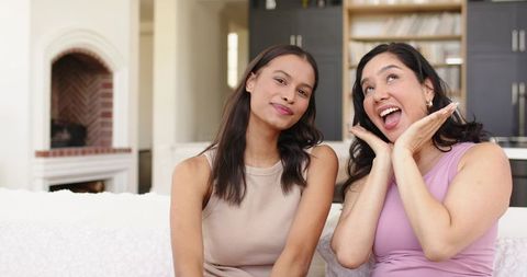 Two friends laughing and smiling on sofa, cozy living room with fireplace mantel