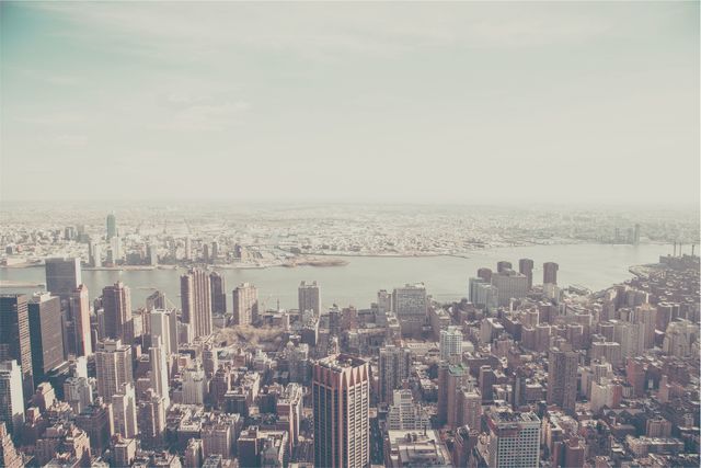 Depicting a sprawling urban skyline with numerous tall skyscrapers and buildings, this image has a hazy atmosphere and overlooks a river separating the dense city. Suitable for use in illustrating topics related to urban development, city living, business districts, environmental studies, and travel brochures. Perfect for backgrounds in advertisements, editorial pieces, and website banners.