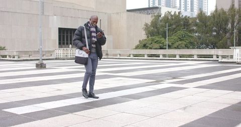 African american man walking across urban plaza checking phone with messenger bag