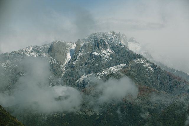 A breathtaking view of a mountain range shrouded in mist, with snow-covered peaks emerging through the fog. Ideal for use in nature-related content, travel blogs, winter holiday promotions, and backgrounds for inspirational posters. It captures the tranquility and majesty of untouched wilderness, making it perfect for emphasizing the beauty of nature and outdoor adventures.