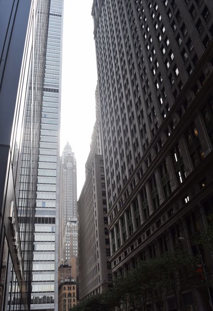Captured from street level, this image features tall glass and stone skyscrapers creating a narrow alleyway. The high-rises stand in contrast to one another, emphasizing modern urban architecture. Suitable for use in articles about city living, urban development, business districts, or architectural designs.