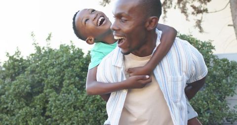 Father giving piggyback to laughing son in backyard, joyful family playtime