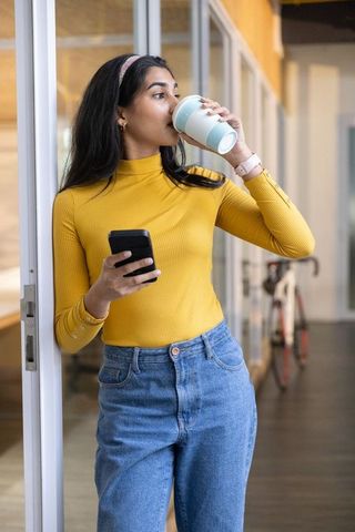 Indian woman drinking coffee while checking phone in modern coworking corridor