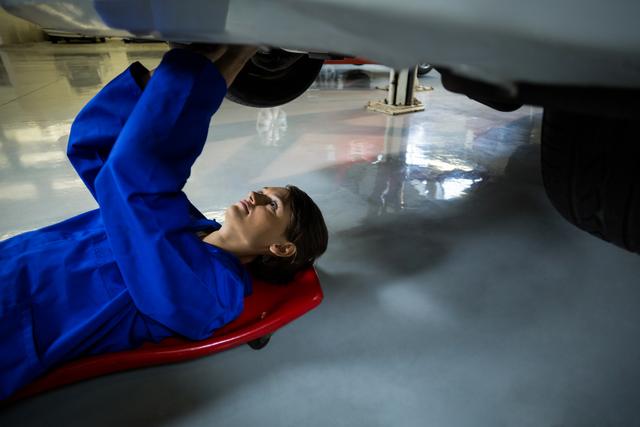 Female mechanic lying on creeper, repairing car in professional garage. Ideal for websites and articles about gender diversity in traditionally male jobs, automotive industry, and skilled trades. Useful for marketing auto repair services, mechanic training programs, and promoting women in STEM fields.