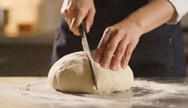 Baker scoring round dough on floured counter with knife for rustic artisan bread