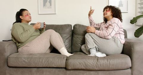 Two friends relaxing on cozy grey sofa wearing loungewear and holding mugs while chatting