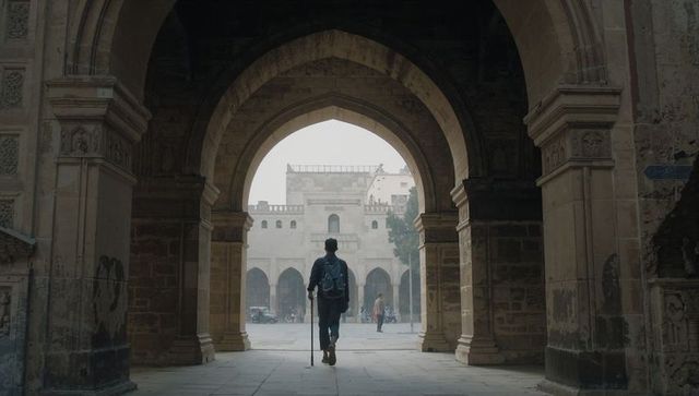 Traveler walking through historic stone archway into sunlit courtyard with backpack and cane