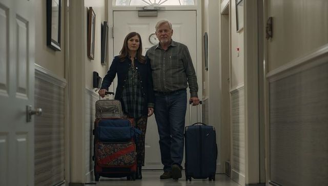 Mature couple leaving family home carrying rolling suitcases through sunlit hallway together