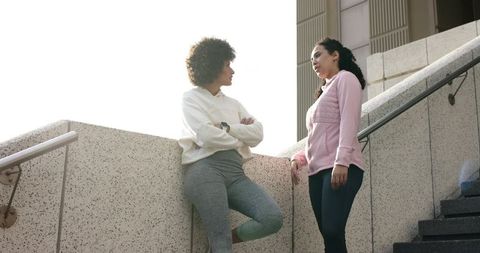 Two women chatting on urban steps in hoodies and leggings during outdoor fitness break