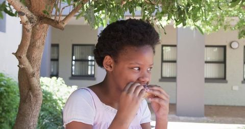 African student eating sandwich under leafy tree on campus courtyard enjoying snack