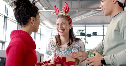Diverse coworkers exchanging holiday gifts in modern office wearing reindeer antlers