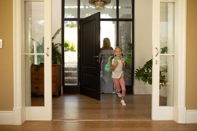 Caucasian mother and daughter arriving at home, mother standing at the open front door while the daughter runs ahead into the hallway wearing a backpack