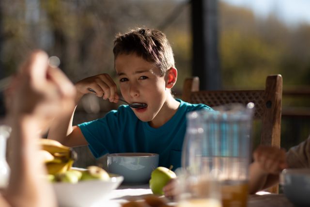 Portrait of a Caucasian boy sitting at a table in the garden enjoying breakfast, eating with a spoon from a bowl looking at camera.. 