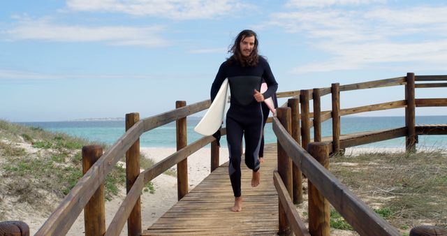 This image depicts a male surfer with long hair carrying a surfboard while walking on a wooden pathway towards the ocean beach. The blue sky with scattered clouds and the tranquil ocean set a serene atmosphere. It can be used for travel blogs, fitness and adventure campaigns, outdoor activities promotions, and coastal lifestyle magazines.