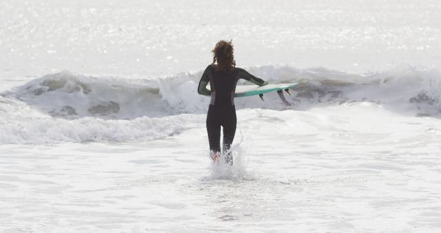Person entering ocean with surfboard underarm, preparing for surfing adventure. Ideal for themes related to surfing, water sports, beach activities, outdoor leisure, summer fun, and adventure.