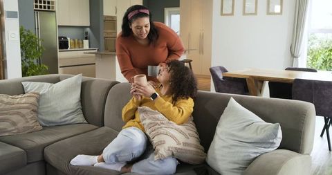 Mother and daughter sharing cozy morning on sofa holding mugs and hugging pillow