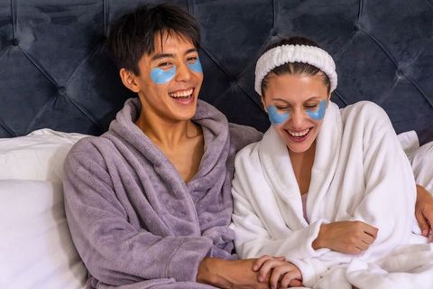 Laughing diverse couple holding hands wearing hydrogel eye patches in plush bathrobes