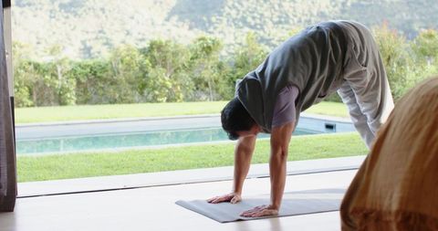 Asian man practicing yoga outdoors by pool through patio door