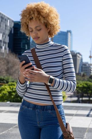 African american woman scrolling smartphone in sunlit urban plaza wearing striped top