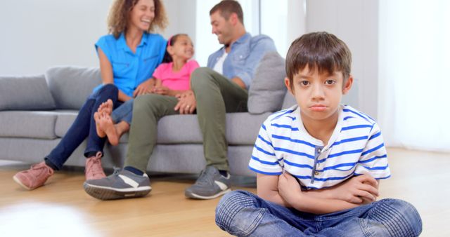 Depicts a young boy feeling upset while sitting on the floor with arms crossed, in clear contrast to the rest of his family who are laughing and enjoying time on the sofa in the background. Useful for themes involving family dynamics, children's emotions, and sibling relationships. Ideal for articles or advertising addressing child psychology, family counseling, and emotional well-being.