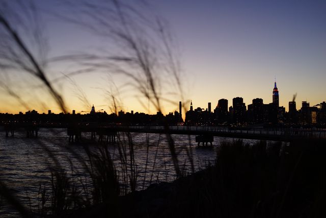 Beautiful city skyline silhouetted against sunset sky, framed by grass in foreground. Water in front enhances tranquil evening ambiance. Perfect for travel blogs, urban photography themes, or serene nature juxtaposed with city life visuals.