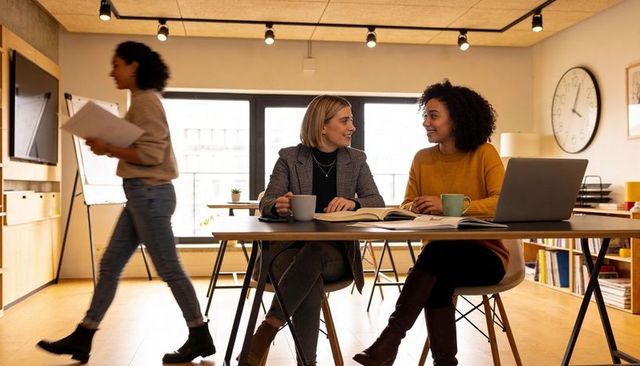 Two female coworkers collaborating at modern office table with laptop, notebooks, coffee