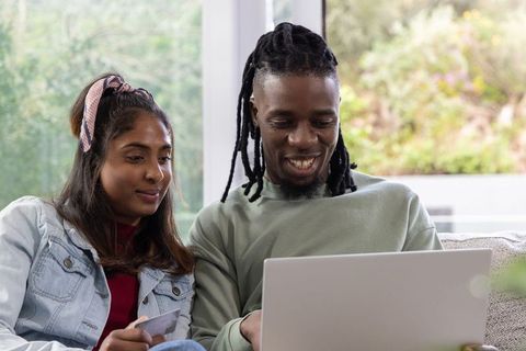 Couple shopping online together on laptop while holding credit card in sunlit living room