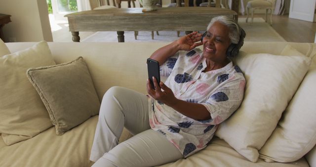 Senior woman enjoying a video call with headphones on smartphone while relaxing on a couch at home. Perfect for illustrating senior wellness, communication with loved ones, and use of modern technology by older adults. Suitable for marketing materials related to tech products, health and lifestyle for seniors, and home comfort furnishings.