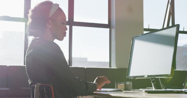 Professionally dressed African American woman working diligently at her computer in modern, sunlit office. Suitable for depictions of workplace diversity, professional environments, women in technology, or career-driven individuals. Could be used in articles or advertisements focused on business, career development, or workplace settings.
