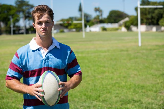 Young rugby player standing on a sunny sports field, holding a rugby ball. Ideal for use in sports-related content, fitness promotions, team sports advertisements, and outdoor activity campaigns.