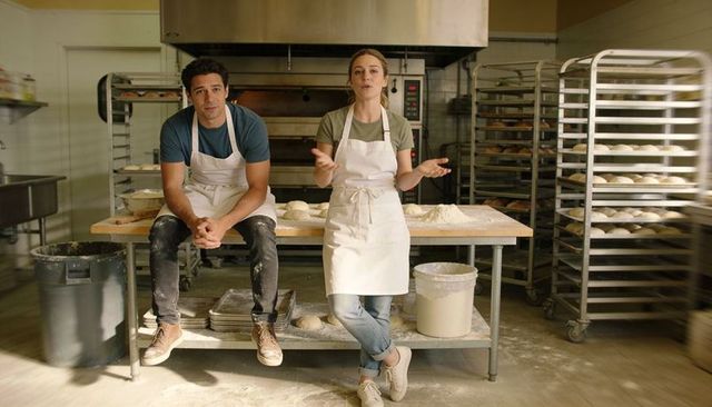 Two bakers talking and kneading dough at wooden worktable with proofing racks and oven