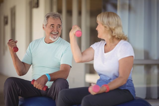 Senior couple lifting dumbbells while sitting on exercise balls on a veranda. Both are smiling and appear to be enjoying their workout. This image can be used for promoting senior fitness programs, healthy aging, active lifestyles, and wellness in retirement communities.