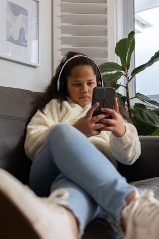 Preteen girl relaxing on sofa with headphones and smartphone, browsing content at home