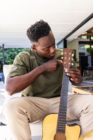 African american man tuning acoustic guitar on covered patio wearing olive tee