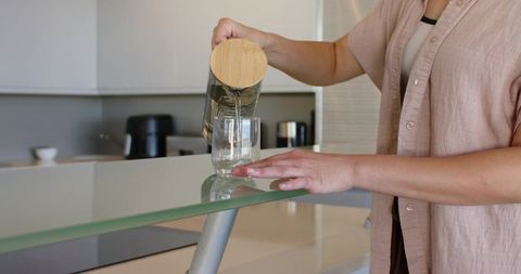 Pouring water from bamboo-lid carafe into glass on glass counter in minimalist kitchen