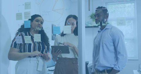 Diverse team collaborating around glass board with sticky notes and tablet for planning