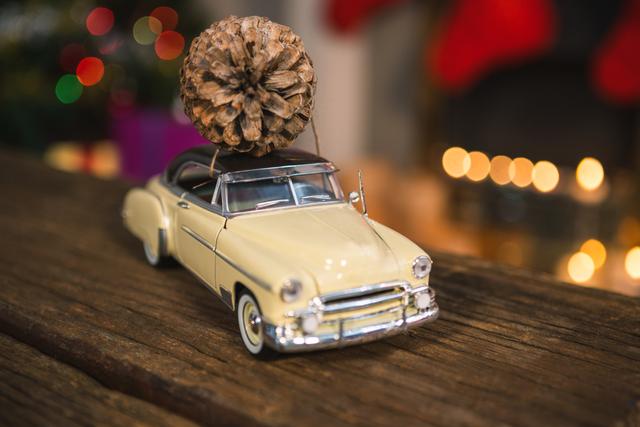 This image features a vintage toy car with a pine cone on its roof, placed on a wooden table. The background is adorned with Christmas lights and festive decorations, creating a cozy holiday atmosphere. Ideal for use in holiday greeting cards, Christmas-themed advertisements, or nostalgic holiday blog posts.