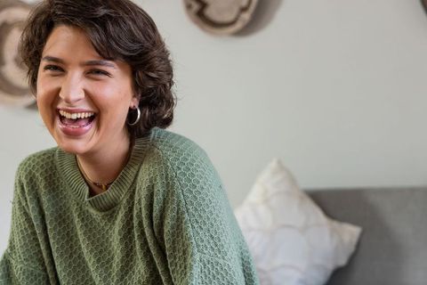 Laughing young woman in green knit sweater sitting on cozy sofa, candid lifestyle portrait
