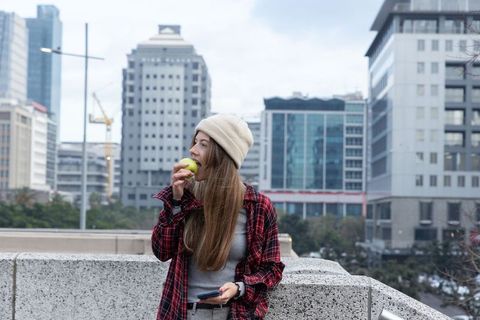 Young woman eating green apple on urban rooftop wearing beanie and holding smartphone
