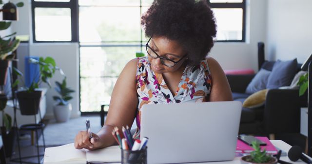 African American woman working at a home office desk, using a laptop, taking notes, and wearing glasses. The modern workspace includes plants and ample daylight. Ideal for illustrating concepts of remote work, productivity, online education, work-life balance, and home office setups.