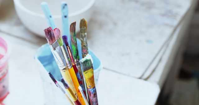 This image shows various paintbrushes standing in a jar, ready for use. The vibrant, multicolored tips indicate they have been recently used for painting. The scene suggests a busy artist's studio, ambiance filled with creativity and inspiration. Ideal image for art supply stores, creative workshops, instructional art manuals, and other artistic-related content.