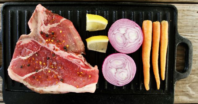 Raw steak seasoned with spices is ready for grilling alongside sliced onions and carrots on a black grill pan. Preparing a balanced meal with protein and vegetables showcases a common home cooking scene.