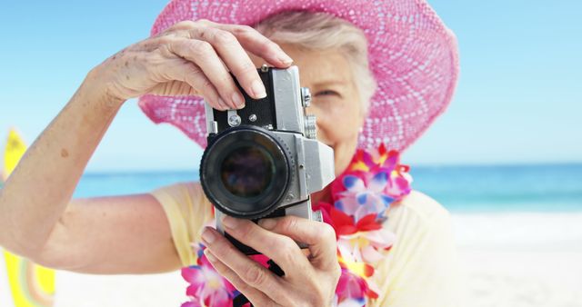 Elderly woman enjoying vacation on a tropical beach while taking photographs with a vintage camera. She wears a pink summer hat and a colorful lei, creating a cheerful and lively image perfect for use in travel brochures, vacation packages, senior lifestyle blogs, and promotional material for photography workshops.