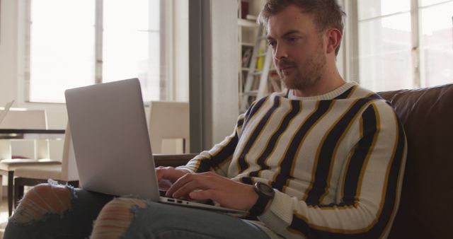 Young man typing on laptop while sitting on couch in home office. Natural light from large windows illuminates the room. He wears a striped sweater and ripped jeans, portraying a casual and modern lifestyle. Perfect for use in articles or blogs about remote work, home office setup, productivity tips, technology use, or modern work-life balance.