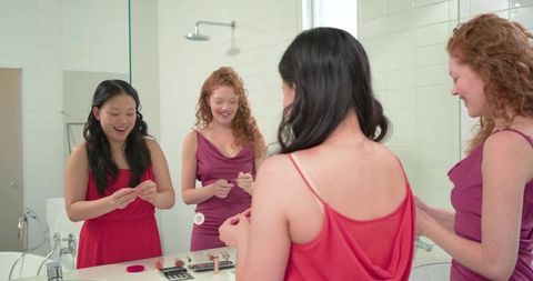 Diverse friends applying makeup at bathroom vanity wearing red and magenta dresses smiling