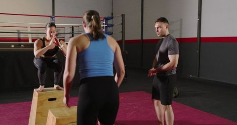 Women performing plyometric box jumps with male coach by boxing ring for gym workout