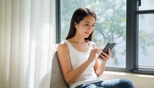 Asian woman using smartphone by sunlit window, casual home lifestyle and mobile browsing