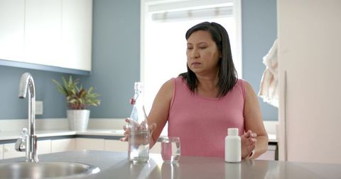 Middle-aged woman pouring water and placing pill bottle on modern kitchen counter