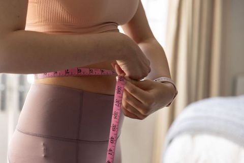 Woman measuring waist with pink tape while wearing mauve leggings and fitness band