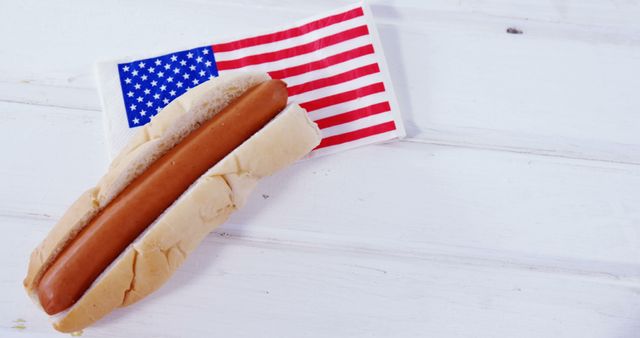 Shows a hot dog in a bun resting on an American flag napkin on a light wooden table. Ideal for themes about American culture, food festivals, national celebrations, barbecues, and summer events.